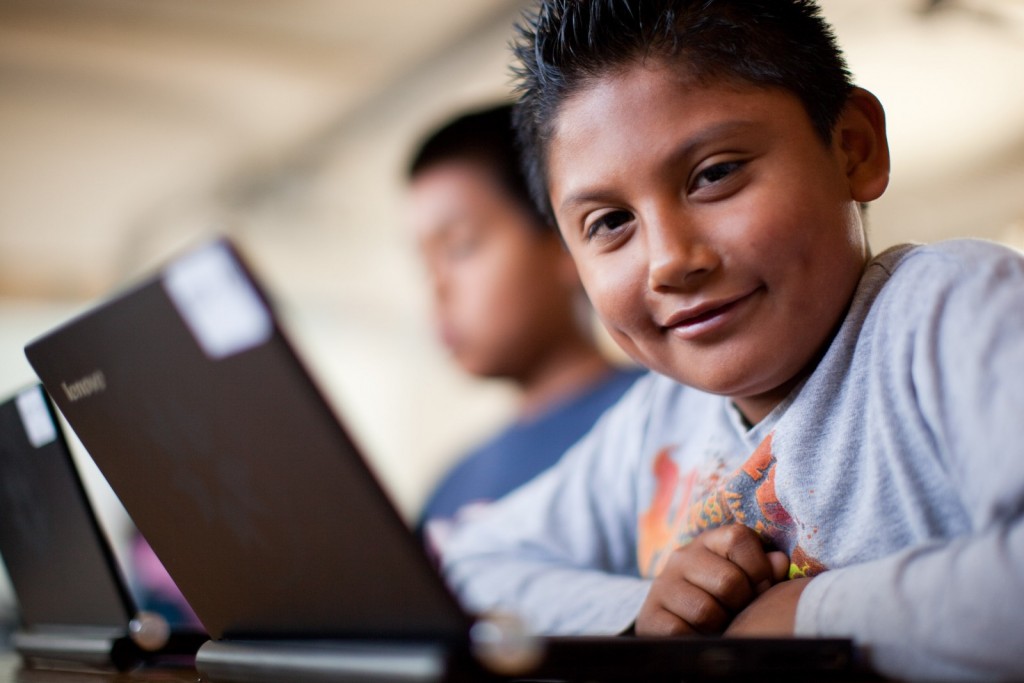 A young boy smiles at the camera while seated at a desk, using a laptop. Another child is seated beside him, also focused on a laptop. Both children appear to be in a classroom setting.