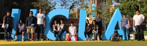 A diverse group of 13 people stand and pose outdoors around large blue letters spelling "UCLA" on a yellow platform. Some are sitting on or leaning against the letters, while others stand beside them. Green trees and a brick building are visible in the background.