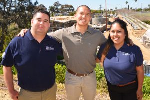 Three people stand side by side, smiling with their arms around each other. They are in front of a construction site with wooden frames and building materials. The two people on the outside are wearing navy shirts with logos, and the person in the center is in a gray polo shirt.