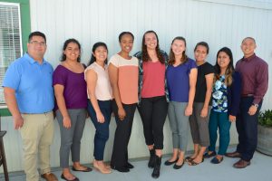 A diverse group of eight women and two men stand close together in front of a white wall, smiling at the camera. They are dressed in a variety of casual to semi-formal outfits, suggesting a relaxed yet professional environment.