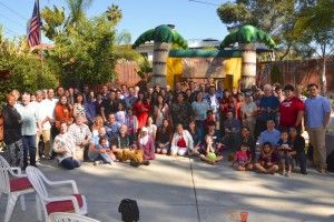 A large group of diverse people gathered outdoors in front of an inflatable palm tree bounce house. The group includes men, women, and children, all smiling and posing for the photo. There are chairs and trees in the background along with various decorations.