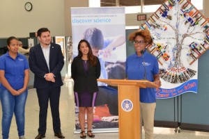 A young woman speaks at a podium labeled with an emblem, while two women and one man stand behind her. The backdrop includes a "discover science" banner and an artistic decoration with a tree and numerous colorful items.