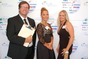 Three people are posing for a photo at an event. The man on the left is wearing a black suit and tie, with glasses, and holding a clipboard. The two women are dressed in formal attire, standing in front of a backdrop with multiple sponsor logos.