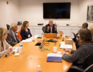 A group of seven people is seated around a large wooden table in a conference room. The person at the head of the table is speaking and gesturing with both hands. Laptops, documents, and water bottles are on the table, and a TV is mounted on the wall behind them.