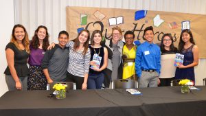 A group of ten people stands in a line, posing for a photo in front of a wall decorated with drawings of books and "Our Heroes" written in large letters. They are smiling and dressed casually. A table with flowers and books labeled "2016" is in front of them.