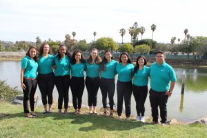 A group of nine people stands side by side on a grassy area in front of a serene lake with trees and palm trees in the background. They are all wearing matching teal polo shirts and black pants, smiling and enjoying a sunny day.