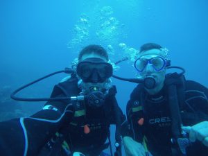 Two scuba divers underwater wear black wetsuits, masks, and breathing apparatus. They are close to each other, with bubbles rising above them. The background is a blue, aquatic environment with some indistinct marine life.