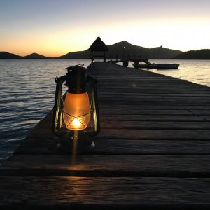 A lit lantern rests on a wooden pier extending into a calm body of water at sunset. The silhouette of a thatched gazebo is visible at the end of the pier, and mountains can be seen in the background against the fading light of the evening sky.