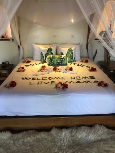 A neatly made bed with white sheets, decorated with green leaves and red flowers spelling out "WELCOME HOME LOVE MOM & DAD." There is a sheer white canopy hanging above the bed and a fluffy beige rug on the floor in front of it.