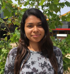 A young woman with long, dark hair stands in front of a lush green plant, wearing a gray floral-patterned top. She smiles slightly at the camera, with a modern building in the background and bright, sunny weather.