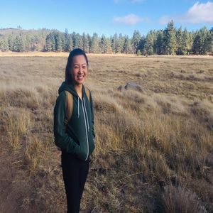 A person in a green hoodie and backpack stands smiling in an open field with tall grasses, surrounded by a forest of pine trees under a blue sky with scattered clouds.