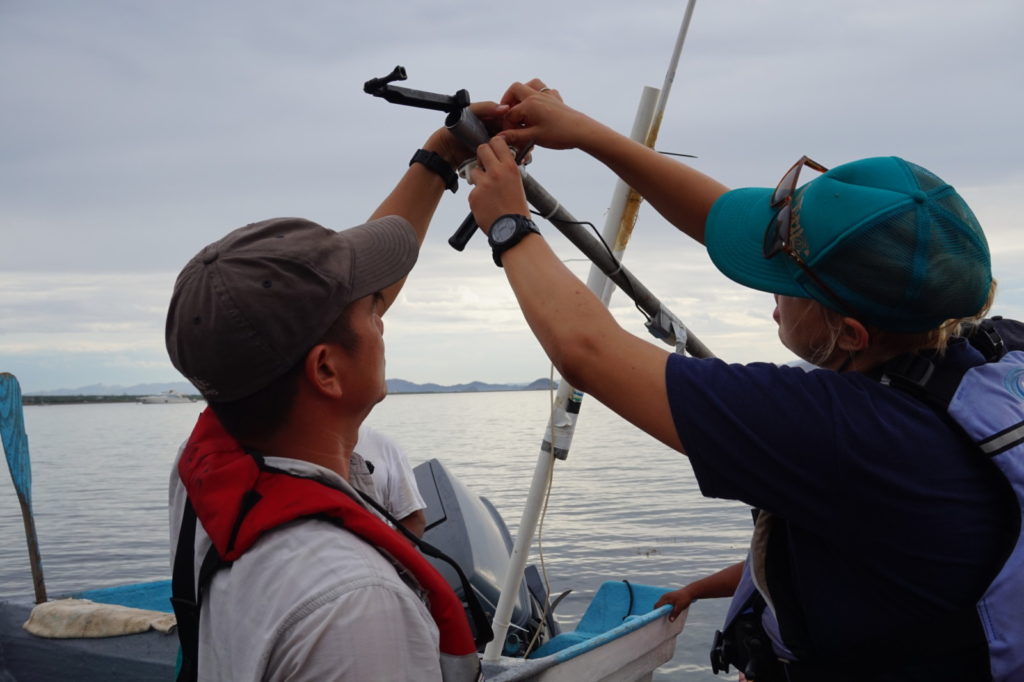 Two people work together on a boat, adjusting equipment mounted on a pole. One person wears a gray cap and a red life jacket while the other wears a teal cap and blue life jacket. The scene is set on a calm body of water with a mountainous horizon under a cloudy sky.