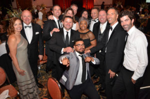 A group of 12 people dressed in formal attire pose together at an indoor event. Most are smiling or laughing, with one person in the front crouching and playfully pointing at the camera. The background reveals tables with decorations, suggesting a festive occasion.