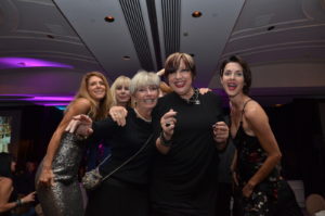 A group of five people, four women and one man dressed in black, pose excitedly at an indoor event with purple lighting. They are smiling, with some pointing towards the camera. The setting has a high ceiling and elegant decor.
