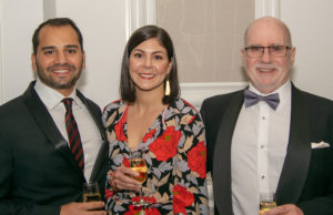 Three people smiling at the camera while holding glasses of champagne. The two individuals on the left are wearing formal attire, with the man in a black suit and striped tie and the woman in a floral dress. The man on the right is wearing a tuxedo with a bow tie.