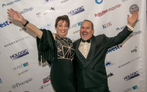 A woman and a man dressed in formal attire stand in front of a backdrop with sponsor logos. Both have their arms raised and are smiling widely. She wears a black and gold dress, while he is in a tuxedo. The backdrop includes logos like "San Diego," "Hologic," and "Kaiser.