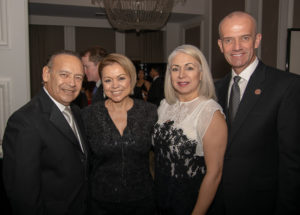 Four people are smiling for the camera at a formal event. The two women in the middle are wearing elegant dresses, while the men on the left and right are dressed in suits. A chandelier is visible in the background.