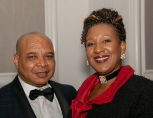 A bald man wearing a black tuxedo and bow tie stands next to a woman with curly hair, wearing a black outfit with a red collar and a jeweled choker. Both are smiling and posing in what appears to be a formal setting.