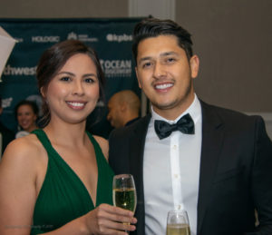 A woman in a green dress and a man in a black tuxedo smile while holding glasses of champagne at an event. A blurred background shows a banner with text and other attendees.