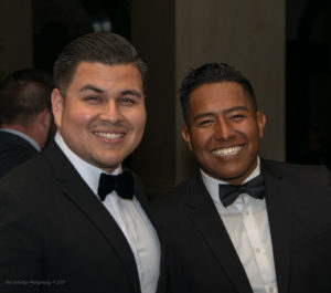 Two men smiling at the camera while dressed in black tuxedos and bow ties. Both have short, dark hair and are standing close together. The background is dimly lit but shows additional people in formal attire.
