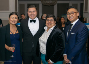 A group of four people dressed in formal attire pose for a photo indoors. Two individuals are wearing suits with ties, one is in a black suit with a bow tie, and the other is in a dark dress holding a drink. They are smiling, with other people in the background.