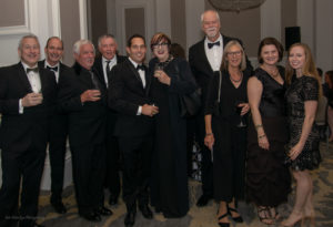 A group of ten people dressed in formal attire are standing together in a room with ornate decor. Some are holding drinks while smiling at the camera. The group includes both men and women, with a mix of dark suits and stylish evening gowns.