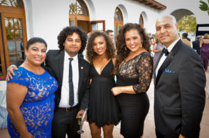 Five people dressed in formal attire at a social event. Three women wear elegant dresses in blue and black, and two men are in suits, with one holding a glass of champagne. They are standing outdoors in front of a building with arched windows.