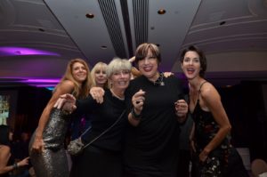 Five women are smiling and posing together at a formal event in a decorated hall with purple lighting. Two women on the left are wearing dresses, the woman in the center is wearing a black sweater and a cross-body bag, and two women on the right are wearing formal black attire.