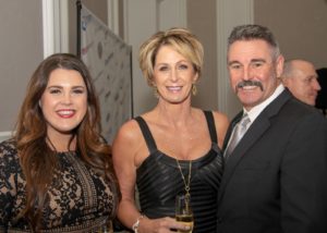 Three people dressed in formal attire smile for a photo at an event. The woman on the left is wearing a black lace dress, the woman in the center is in a black dress with a necklace, and the man on the right is in a suit and tie with a mustache.