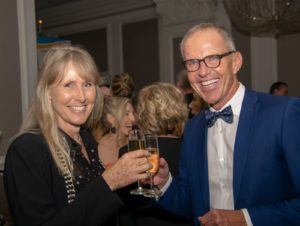 A woman and a man, both dressed formally, smile and toast with champagne glasses at an indoor event. The woman has long blonde hair and wears a black outfit with a beaded necklace. The man, wearing glasses and a blue suit with a bow tie, stands beside her.