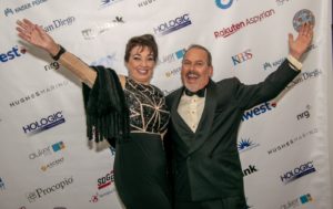 A man and a woman dressed in formal attire are standing together with their arms raised in celebration. They are posing in front of a backdrop filled with various sponsor logos. The woman is wearing a black and gold dress, while the man is in a black tuxedo.