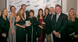 A group of people pose together at a formal event. They are dressed in evening attire, with women in dresses and men in suits. Some hold drinks and papers. A banner behind them displays various logos and sponsors. Everyone is smiling and facing the camera.