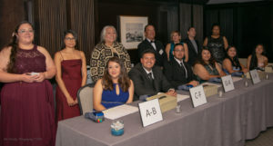 A group of people, dressed formally, stand and sit behind a long table with name tags and boxes marked A-B, C-F, G-H, and L-N, likely organizing paperwork or check-ins. They smile towards the camera in an indoor, professional setting.
