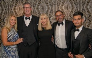Five formally dressed people stand together posing for a photo. The women wear elegant dresses and the men wear tuxedos with bow ties. They are in front of an ornate, patterned backdrop.