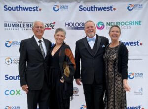 Four people dressed in formal attire are posing together and smiling in front of a step-and-repeat banner featuring logos of various sponsors including Ocean Discovery, Southwest, and others. The group appears to be attending a special event or gala.