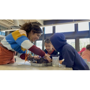 A woman in a colorful sweater is helping two young children with a craft project at a table. The children are focused on the activity, one wearing a hoodie. The setting appears to be a bright, modern classroom or art studio with large windows in the background.