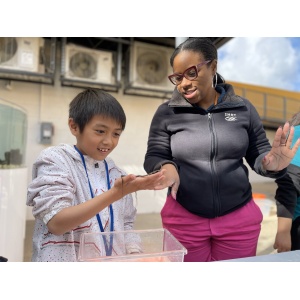 A child and an adult are engaged in a science-related activity outdoors. The child is holding a small object, looking excited. The adult is wearing glasses and smiling while gesturing towards the object in the child’s hand. They are standing next to a clear container.