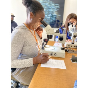 A person with a big smile looks into a microscope while holding a pen and filling out a worksheet in a laboratory setting. Another person is observing a sample through a microscope in the background.