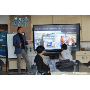 A person in a blue blazer standing next to a large screen, giving a presentation to students seated in a classroom. 