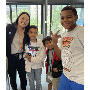 A woman and three children are smiling and posing indoors. One child is making a peace sign. The woman and children wear name tags. The background shows windows and plants outside. The group appears happy and cheerful.