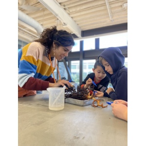 A woman in a multi-colored sweater conducting a science experiment on a table with two students.