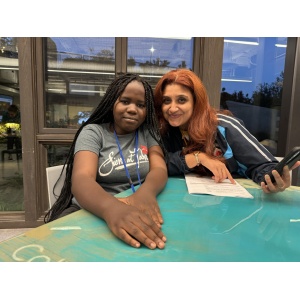 Two women are seated at a table. The younger woman on the left, with long braided hair wearing a gray shirt, and the older woman on the right with reddish hair holding a phone and smiling. A window with a nighttime outdoor view is in the background.