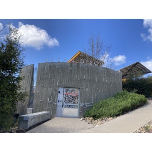 The image shows the entrance to a building with "Ocean Discovery" sign above a curved, textured concrete wall. The door features intricate metal designs. The structure is surrounded by greenery, and the sky is clear with a few clouds.