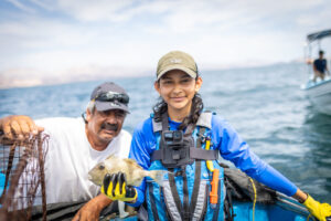 Two individuals on a boat on the ocean. One individual is a man wearing a white shirt with a hat smiling, the other indidivual is female student holding a fish with a camera straped to her chest wearing yellow gloves and a blue long sleeve. 