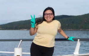 Student with dark hair, wearing glasses, and surgical gloves on a boat in the ocean holding to a wooden ship railing 