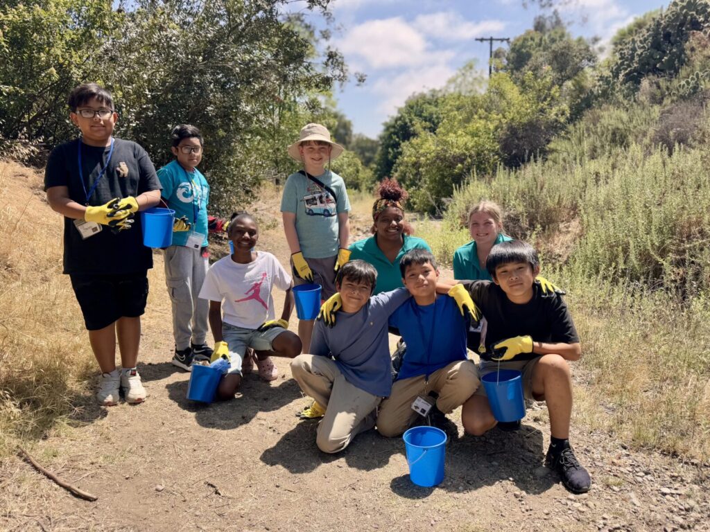 A group of diverse students with two instructors outside in the canyon on a sunny day wearing yellow gloves and holding blue buckets.
