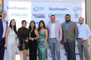 8 individuals of diverse background standing infront of a step and repeat with Southwest Airlines, Ocean Discovery, the Bumble Bee Seafoods Company, Hologic, Neurocrine and Sycuan logo visible, all individuals are smiling